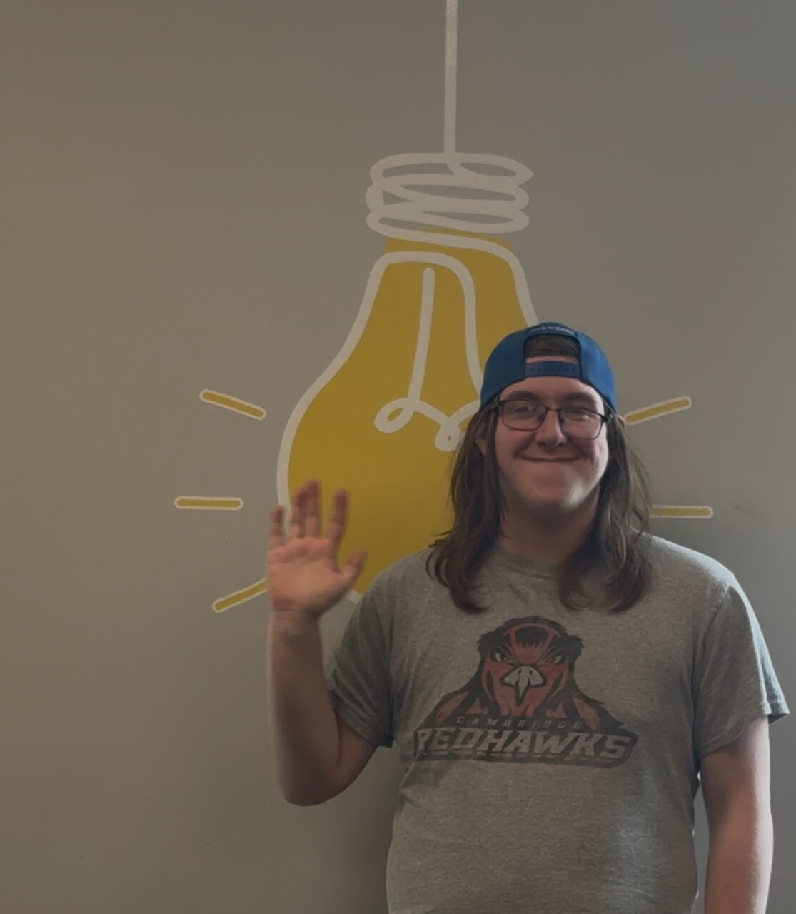 a young white man with a backwards cap waving in front of a giant lightbulb mural on the wall